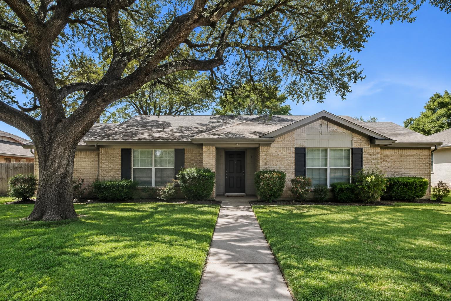 A modest single-story home in San Antonio with a green lawn, a live oak, and a walkway to the front door